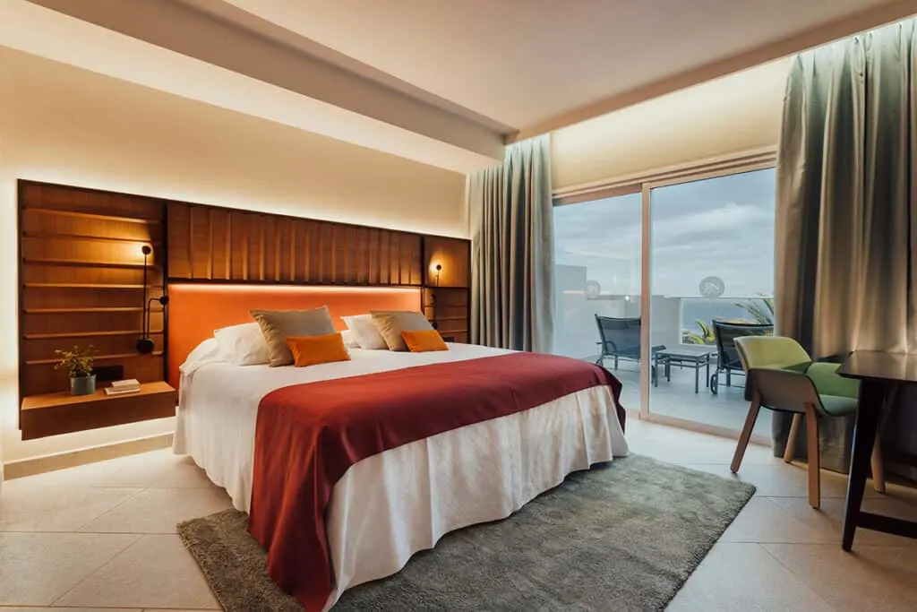 Find the best beachfront hotels Costa Adeje can provide, interior of hotel room with large bed and soft pillows next to writing desk area and large sliding glass doors leading to balcony