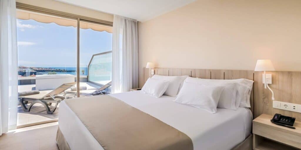 Try out the best beach resorts in Tenerife, hotel room interior with large bed decked out in white and beige color scheme with wide glass sliding doors leading out onto balcony