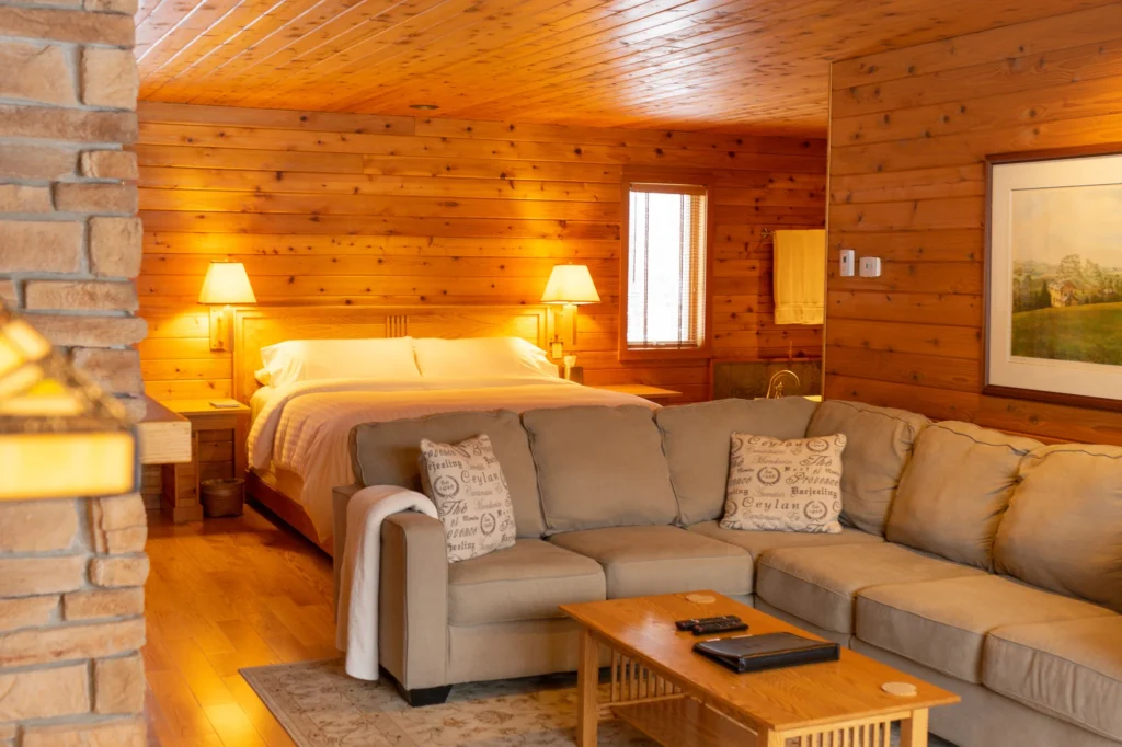 Find your new favourite adults-only resorts in Wisconsin, living room of low-ceilinged wood cabin with large corner sofa and wooden coffee table in foreground and large bed with bright lamps behind