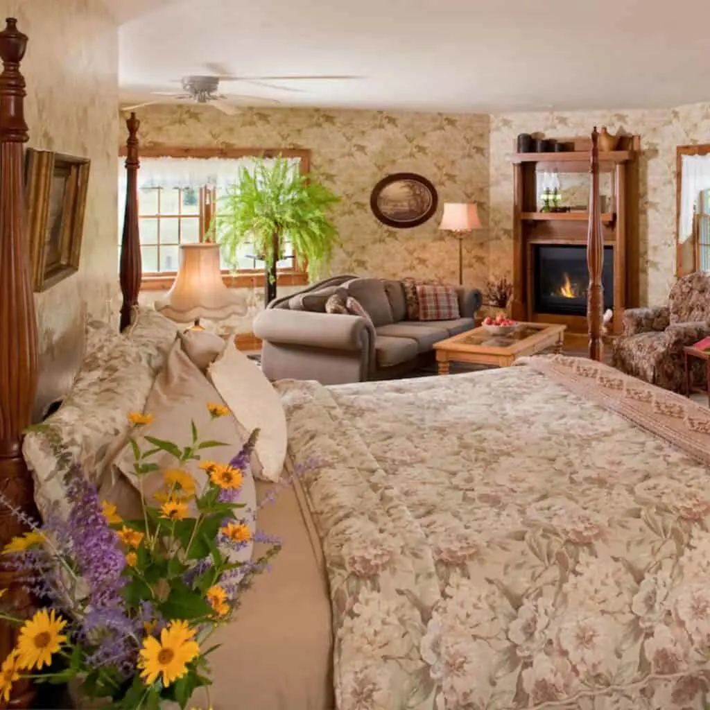 a classic bedroom with brown tones, flowers on the bedside table, a queen bed, couches, lamp and a window