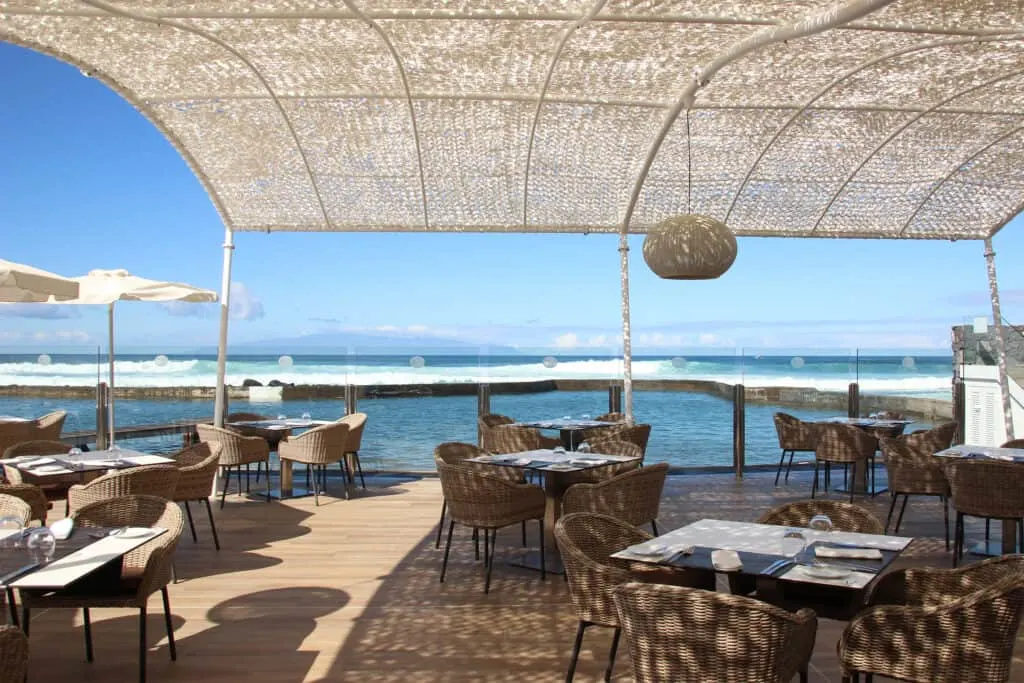 Don't forget to try the beachfront hotels Los Cristianos has to offer, outdoor dining area with sets of tables and chairs sitting on a wooden deck shaded by a light roof with a view of the wide open sea under a clear blue sky