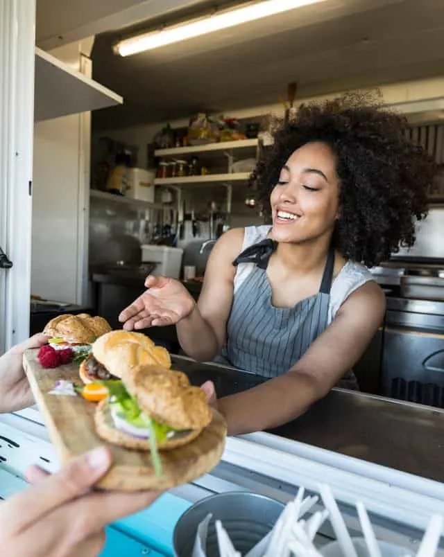 smiling person in restaurant being handed a wooden tray on which are arranged a selection of three burgers