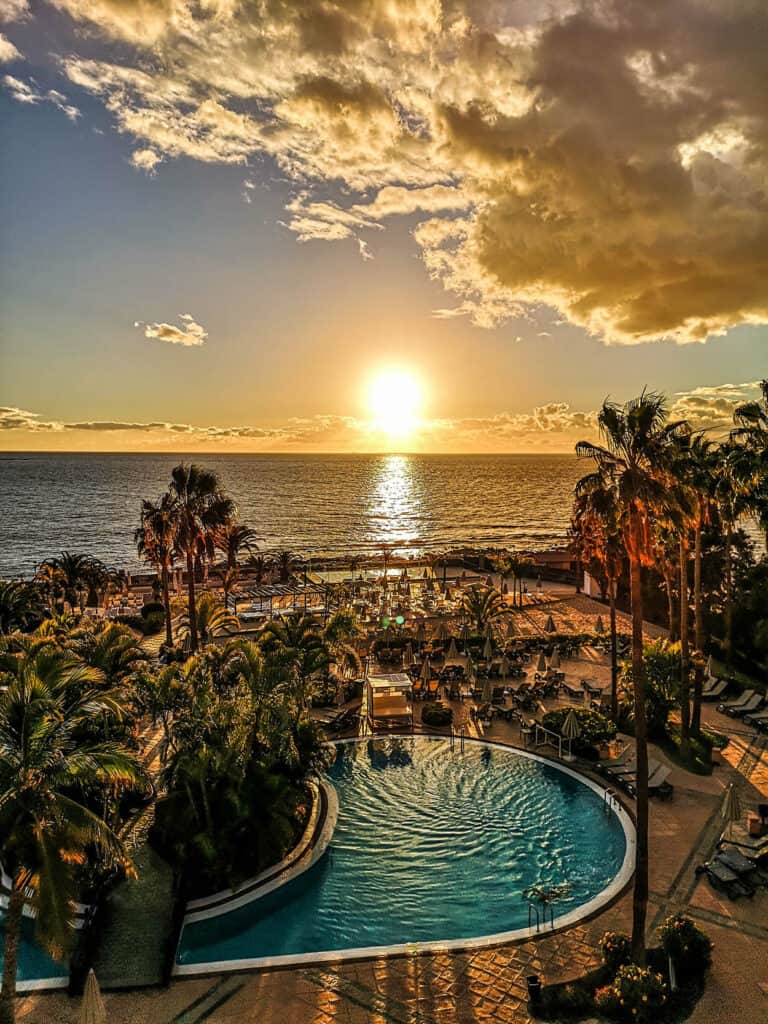 Stay at some of the best Tenerife beach resorts, aerial view of large outdoor pool and beach area with palm trees and sun loungers all facing the calm sea with the sun setting in on the horizon