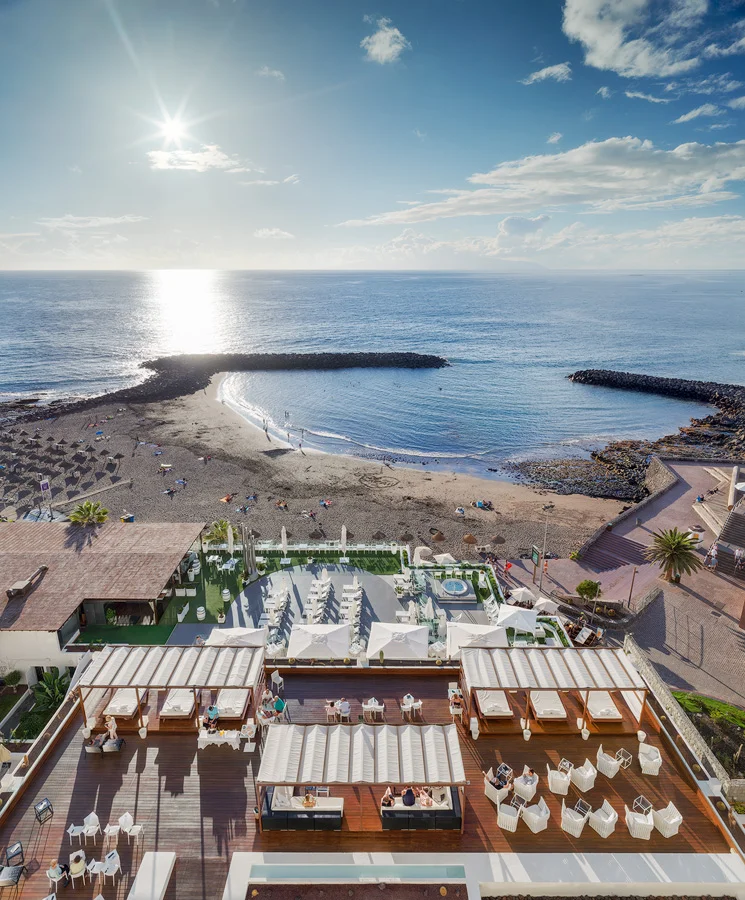 Try out some Tenerife beach apartments on your next vacation, aerial view of hotel outdoor area with sun loungers and covered deck chairs in front of sandy beach area with calm sea waves lapping at the shore