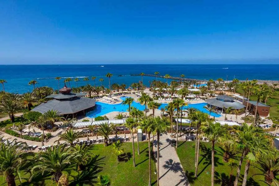 Enjoy these beach hotels in Tenerife, aerial view of carefully organized outdoor hotel pool area with palm trees and pools next to large shaded huts with the vast blue sea behind