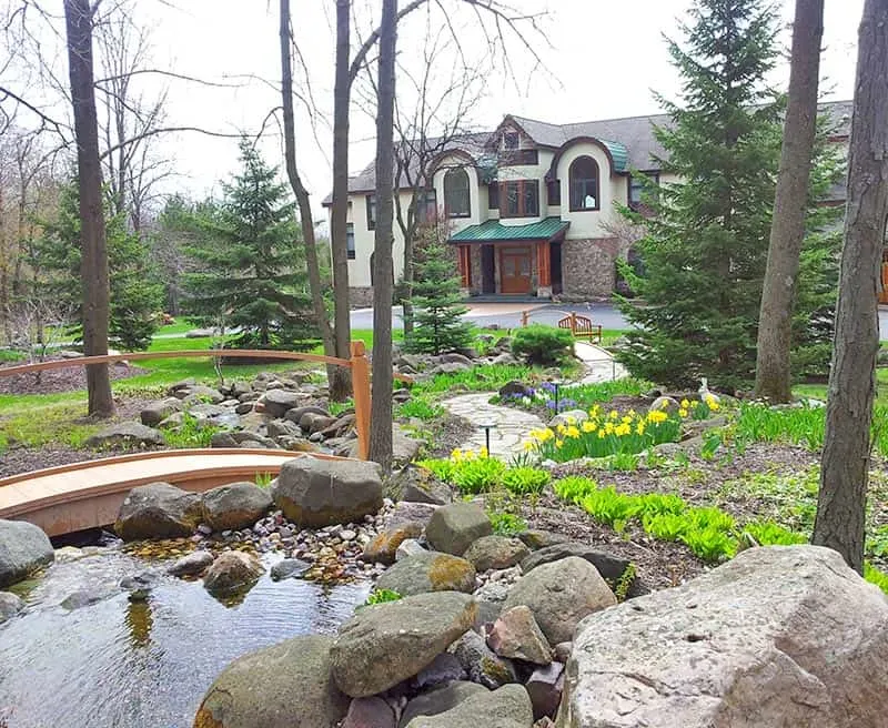 Try out some of these adult vacation ideas in Wisconsin, large lodge with elaborate front garden area featuring pond with rockery and wooden bridge leading into a stone pathway winding through the tall trees and plants