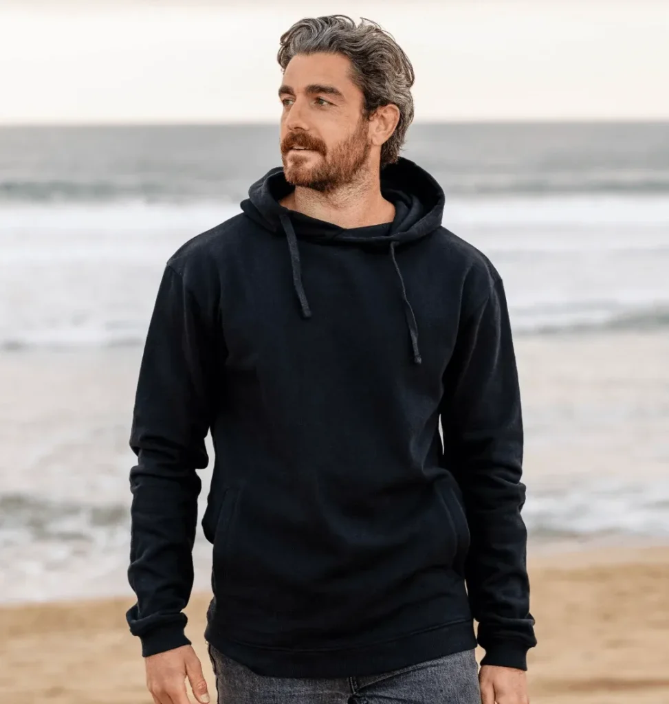 sustainable clothing brands with recycling programs, white man wearing black hood and black jeans walking along a sandy beach