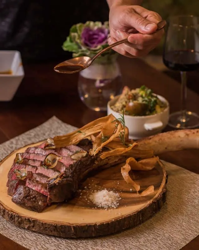 large steak on the bone sitting on a wooden plate with a side dish and glass of water nearby on top of a wooden table