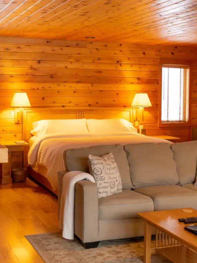 Find your new favourite adults-only resorts in Wisconsin, living room of low-ceilinged wood cabin with large corner sofa and wooden coffee table in foreground and large bed with bright lamps behind