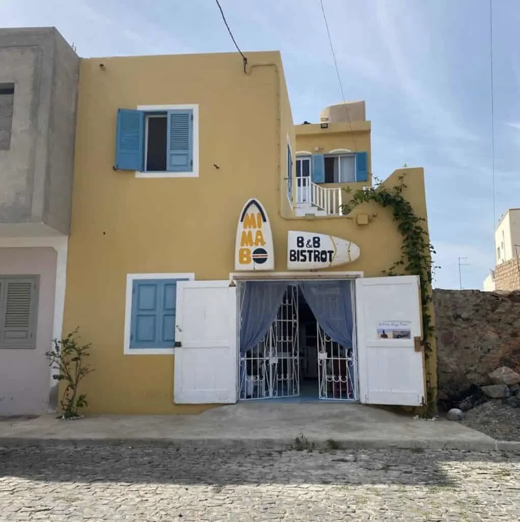 affordable hotels in Boavista Cape Verde,, exterior view of yellow boxy building with blue shutters, doors are open and sign above doors reads Mi Ma Bo B&B