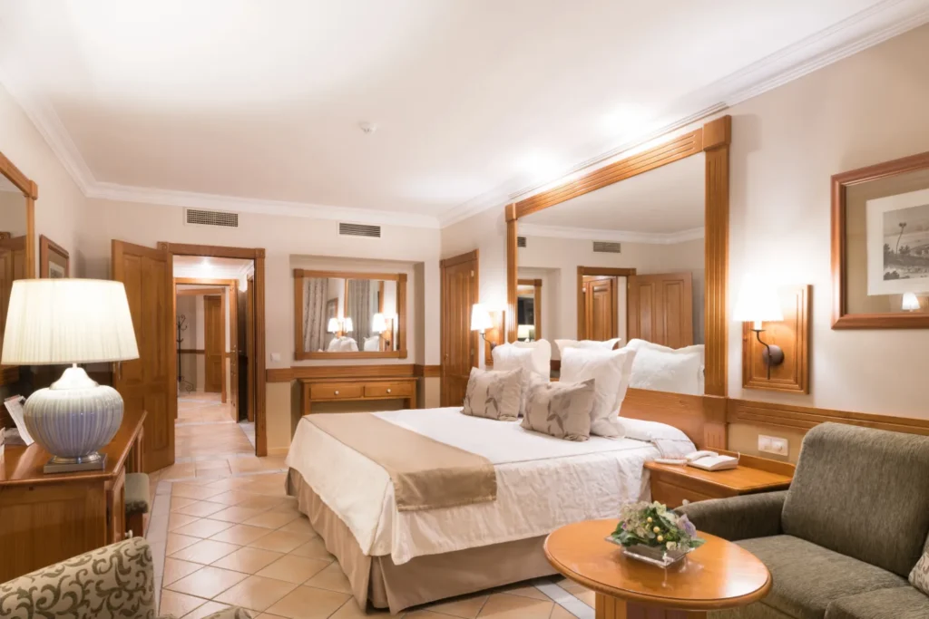 Enjoy some of the best family holidays in Tenerife, hotel suite bedroom with large bed beneath large mirror, room also features a vanity, desk, chair and a couch, room to door is open showing hallway to rest of suite