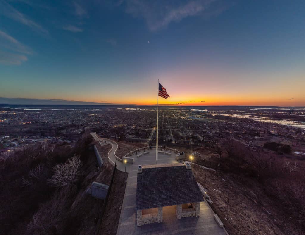best things to do in La Crosse Wisconsin, Aerial view of the top of a hill on which stands a single stone hut and a tall flagpole with a flag of the USA flying high in the breeze with a view out across a vast valley with many built up areas of houses and other buildings all under a wide open sky at sunset