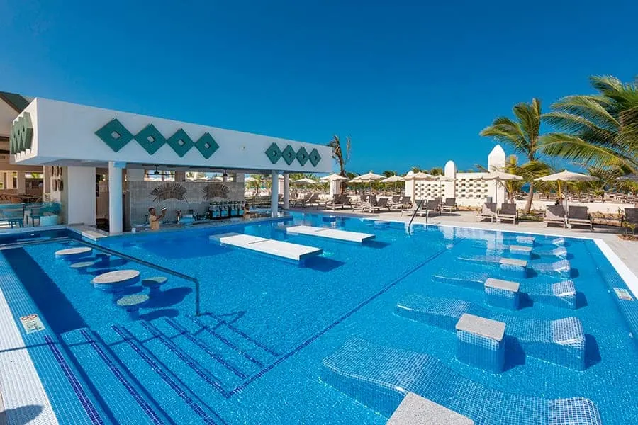 Outdoor swimming pool with submerged tiled benches and stone seating with a bar area to one side and some palm trees on the other all under a clear blue sky