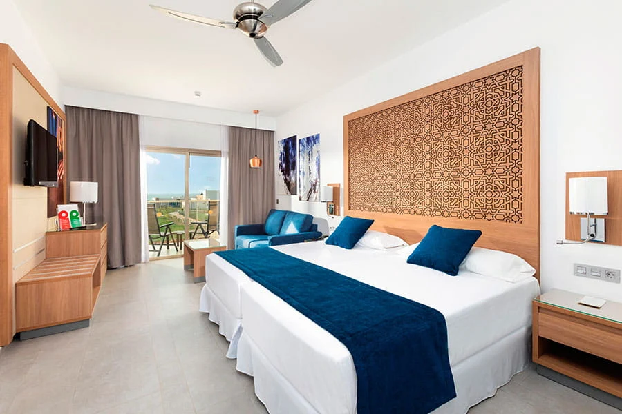 luxury hotels in Boa Vista Cape Verde, Interior of hotel room with neatly made up twin beds and a large wooden headboard with intricate black design with balcony area visible at the rear