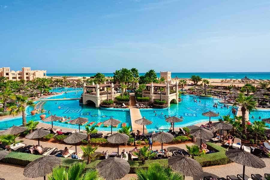best Boa Vista Cape Verde resorts, Aerial view of a large pool and sunbathing area of a hotel with a central island area covered in ornate buildings and green trees connected by four bridges to the rest of the land and the sea visible behind