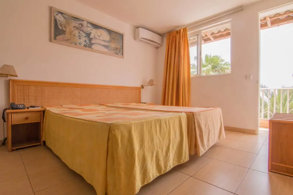 Interior of hotel room with two twin beds next to each other and a bedside table with a phone and a lamp all under a picture of some modern art