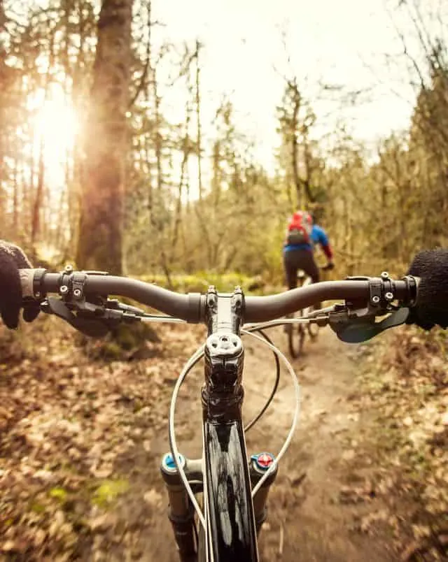 Where to go for Door County biking, POV view from person on bike looking over the handlebars at another person on a bike in bright clothing as they ride along a dirt path through forest in the low evening sun