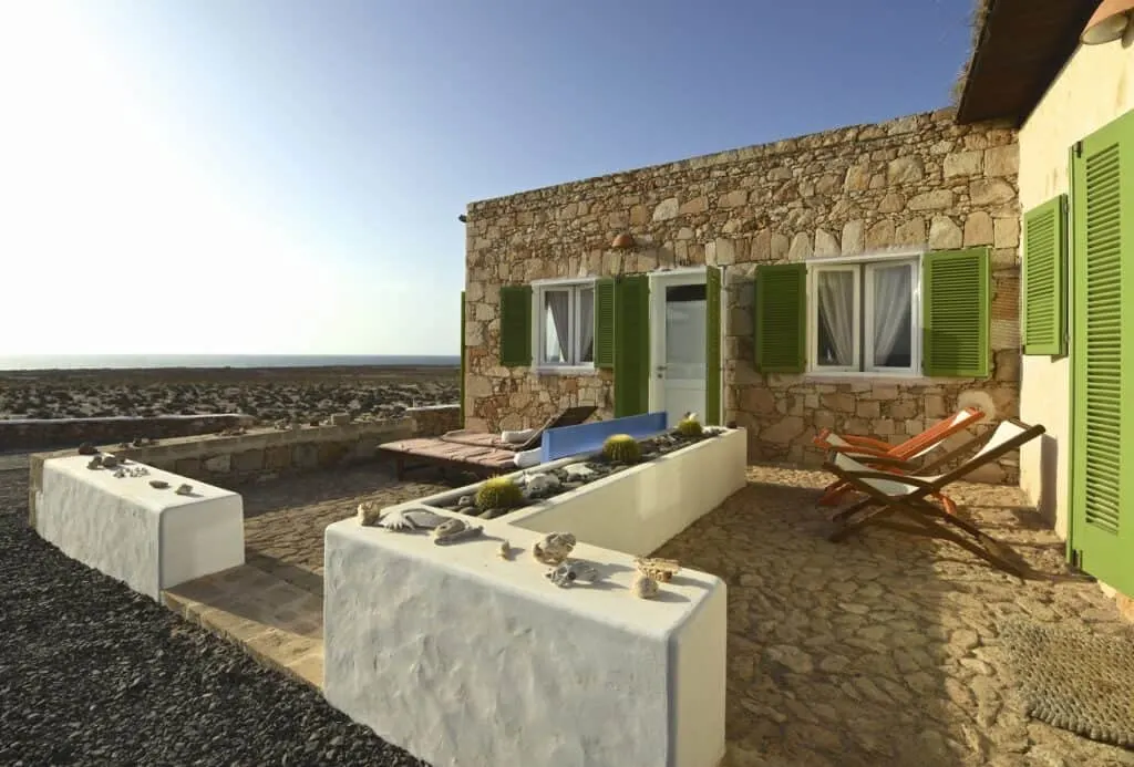 Outdoor sunbathing area with wooden deck chairs and low stone walls next to a single storey stone building under a clear blue sky