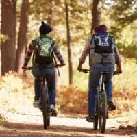 Two people with backpacks and outdoor clothes on bikes having a conversation whilst traveling through forest on a dirt path in bright sunlight in wisconsin