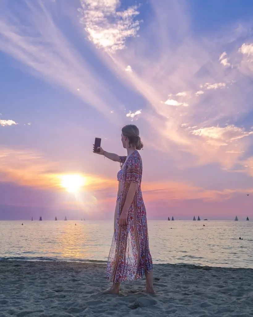 a travel blogger using her phone for a selfie on the beach on a sunset