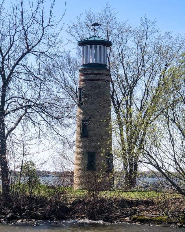 Best places to visit in Oshkosh Wisconsin, Stone lighthouse with three windows in the side standing in a patch of grass surrounded by a handful of trees with no leaves under a bright blue sky
