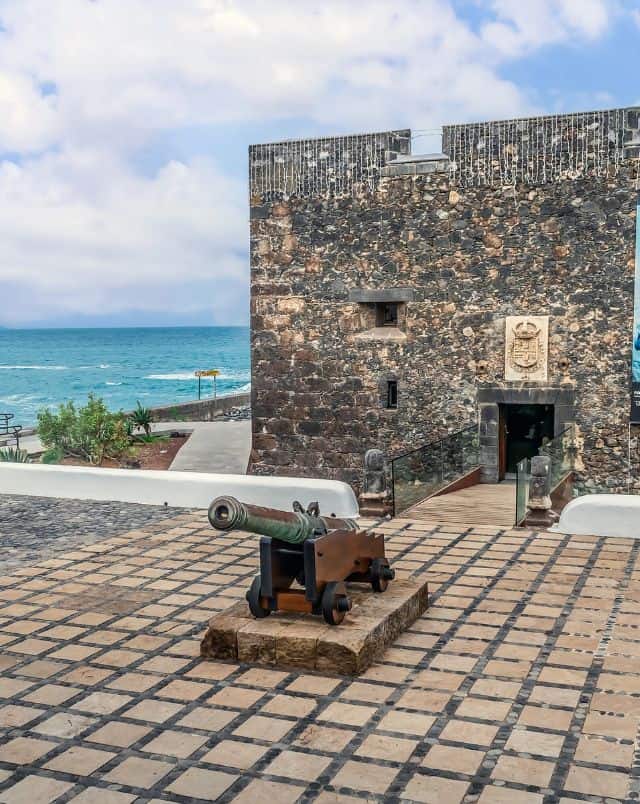 Top Puerto de la Cruz activities, Single cannon sitting on stone tiles outside of a square stone building with small drawbridge acces to the front door and blue sea behind under a blue sky with clouds