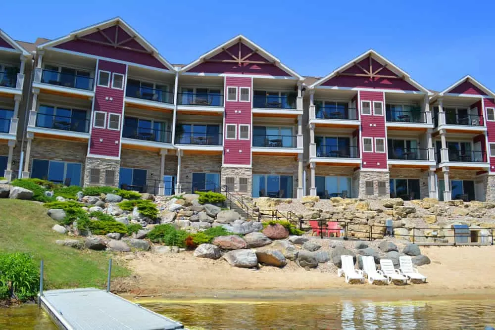 Wisconsin Dells winter hotels, exterior of resort facing water with red and stone romantic Wisconsin Dells hotels, stairs from resort lead down to sandy beach area with white beach chairs