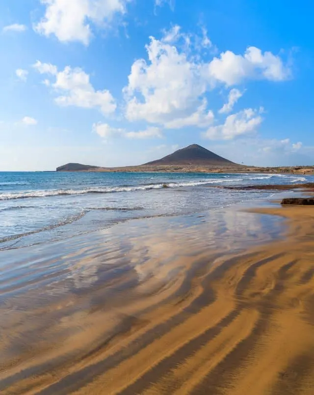 Top beaches South Tenerife has to offer, Clear sea waters lapping lazily on the shores of a yellow sandy beach with a tall pointed hill in the distance all under a bright blue sky with some white fluffy clouds