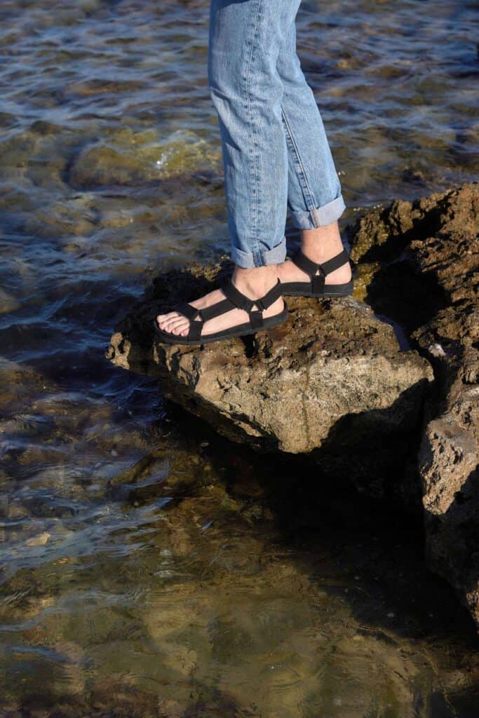 vegan sandals for men and women, bottom half of person standing on rock above water wearing black sandals