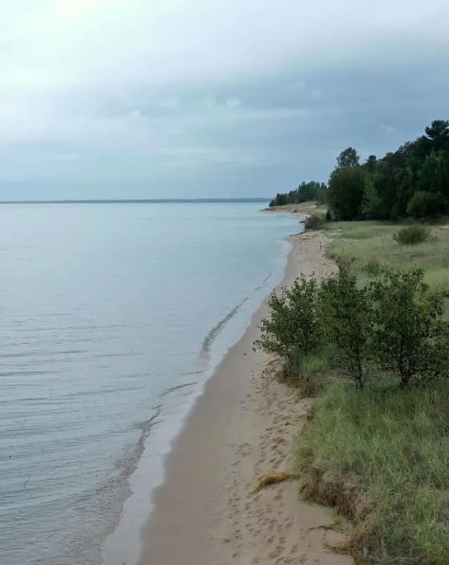 best sandy beaches in Door County, View looking along a long and narrow sandy beach with small waves lapping at the shore and green trees and grass behind all under a grey cloudy sky