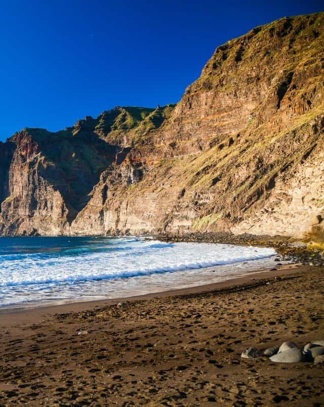 Amazing South Tenerife beaches to visit, Tall rocky cliffs with green peaks standing next to the shores of a sandy beach with white surf lapping at the shore under a clear azure blue sky