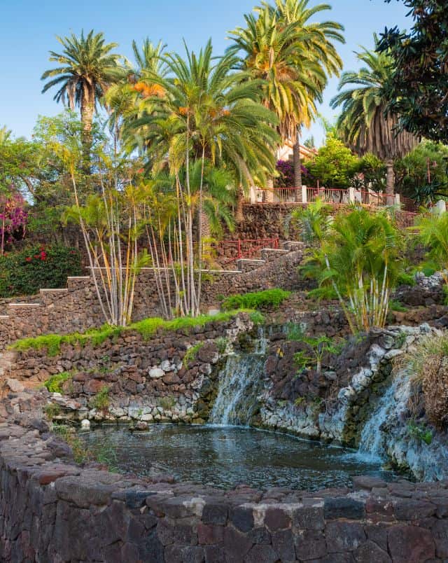 things to see in Puerto de la Cruz, Outdoor pool of water in tiered rocky area with green palm trees placed behind and nearby with some stone steps leading up to a balcony area all under a clear blue sky
