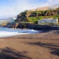 places to visit in Puerto de la Cruz, View of coast with sandy beach in the foreground and road leading over a stone bridge behind next to tall rocky hill with large apartment buildings on top all under a blue sky with white wispy clouds