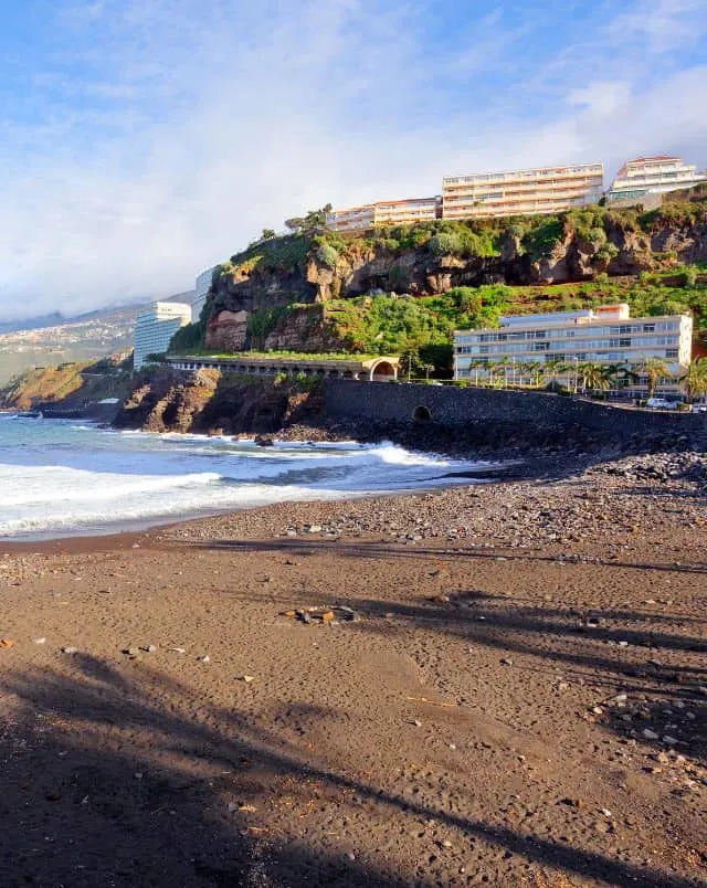 places to visit in Puerto de la Cruz, View of coast with sandy beach in the foreground and road leading over a stone bridge behind next to tall rocky hill with large apartment buildings on top all under a blue sky with white wispy clouds