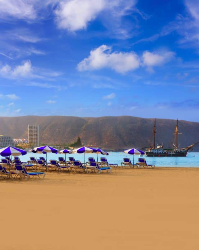 Beautiful beaches in South Tenerife, Yellow sandy beach on which sit lines of blue deck chairs with accompanying blue and white beach umbrellas with the sea behind in which sits a sailing boat with a bank of tall rolling hills behind all under a bright blue sky with some wispy white clouds