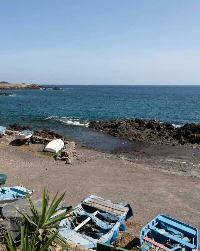 Relaxing Tenerife South beaches, Grey sandy beach with small rowing boats lined up on the shore in the foreground and dark rocks behind on which falls the small waves from the sea which spreads out behind to the horizon all under a subdued blue grey sky
