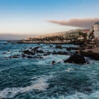 Amazing things to do in Puerto de la Cruz, View of coastline with waves crashing onto dark rocks with large buildings in the distance under a blue sky with some grey clouds