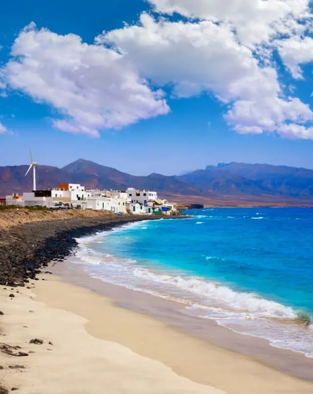 things to do near Puerto de la Cruz, View along the coast with sandy beach in the foreground and buildings further along with large rolling hills and mountains in the distance all next to the vibrant blue waters of the sea and all underneath a wide open blue sky with some thick white clouds