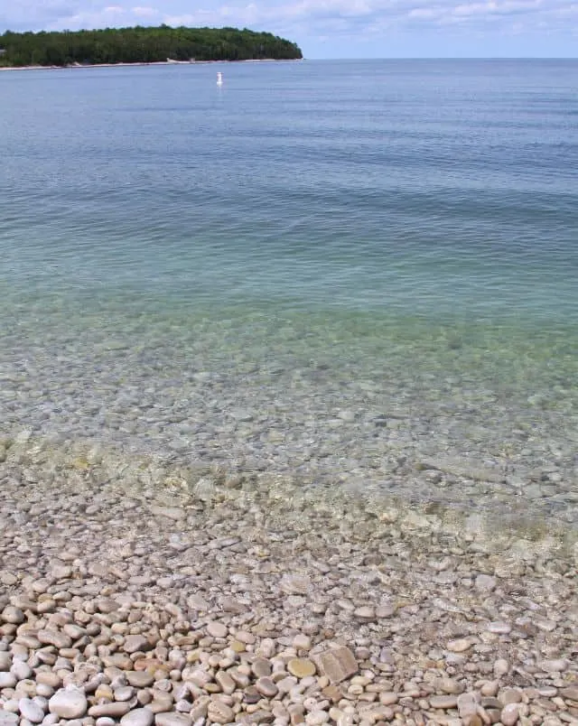 Schoolhouse Beach in Washington Island, View of stone pebble beach with clear turquoise seawater sitting calmly with tree-covered islet area in the background