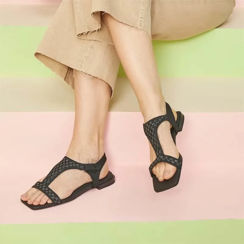 comfortable vegan sandals, crossed legs of someone wearing tan capri pants and black sandals sitting on a pastel pink and green bench