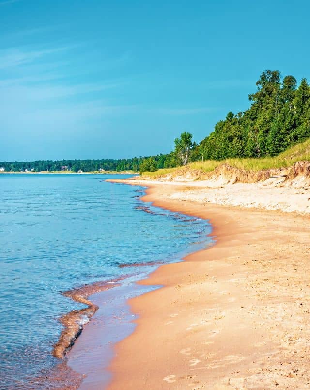 Top Door County Wisconsin beaches, View looking along a sandy beach with small waves lapping at the yellow sand and lush green trees lining the coast all the way along under a bright blue sky