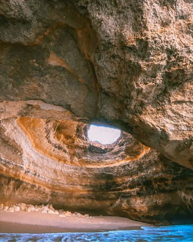 View from inside cave with small sandy area sitting next to the white surf of the ocean with a large rock formation above with a circular hole through which can be seen the sky