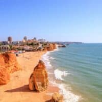 where to go in Algarve Portugal, View of coast with sandy beach and rock formations next to white surf and the wide open sea with a large urban area behind all under a clear blue sky