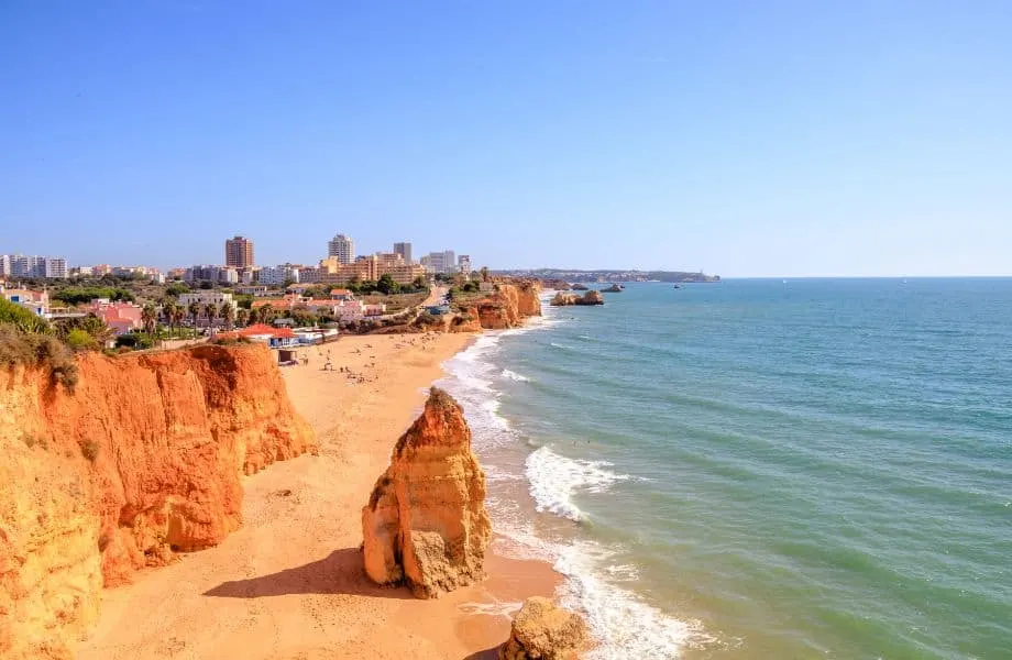 where to go in Algarve Portugal in October, View of coast with sandy beach and rock formations next to white surf and the wide open sea with a large urban area behind all under a clear blue sky