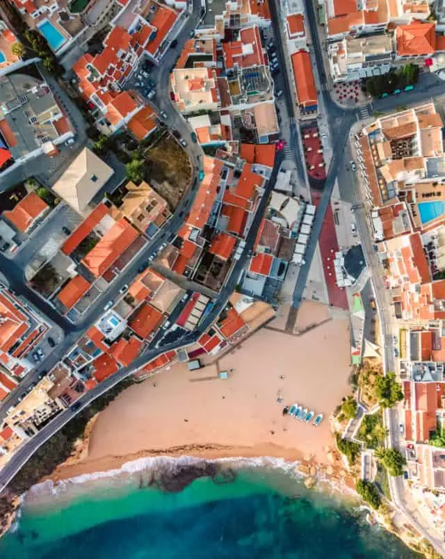 best beach towns in Algarve for a day trip, Aerial view of small sandy beach next to the turquoise sea surrounded by densely built up urban area full of buildings with terracotta rooftops