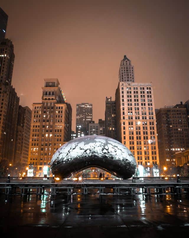 winter getaways from Milwaukee, Wide view of the Chicago Bean sitting in an open outdoor square covered in a light layer of recent rain with tall skyscrapers behind under a dark cloudy sky at night