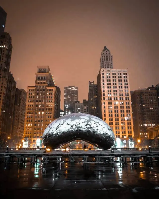 winter getaways from Milwaukee, Wide view of the Chicago Bean sitting in an open outdoor square covered in a light layer of recent rain with tall skyscrapers behind under a dark cloudy sky at night