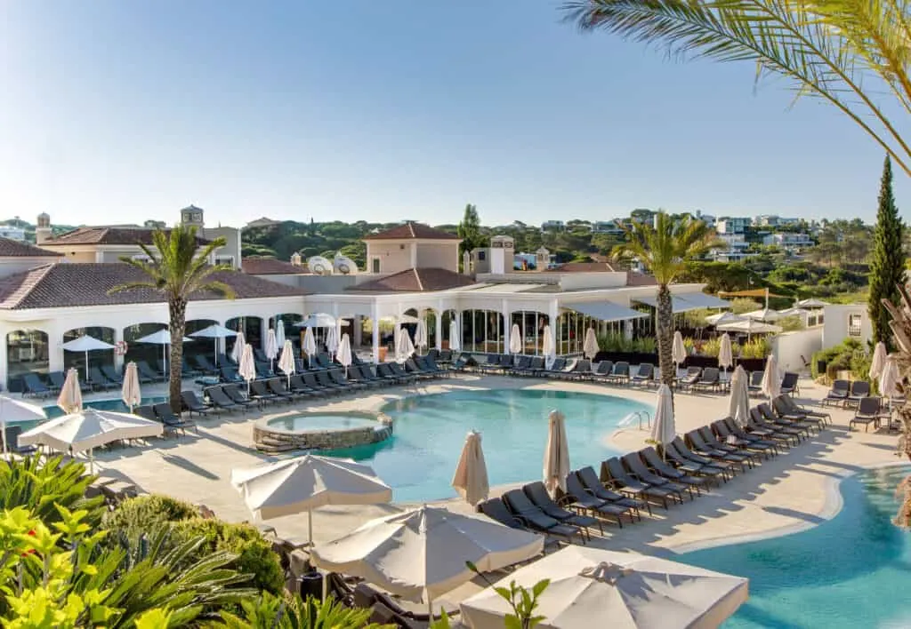 Enjoy some of the best 5-star family hotels Algarve, view of large outdoor pool area with lines of sun loungers lining the edge as well as sporadic palm trees and rock-walled smaller pool next to the main pool