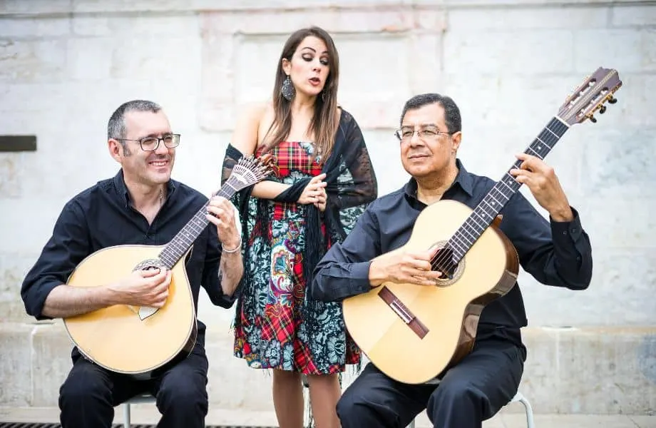 Musicians playing in the street with two men in black shirts and glasses holding stringed guitar instruments while a lady in a patterned dress and black shawl sings between them