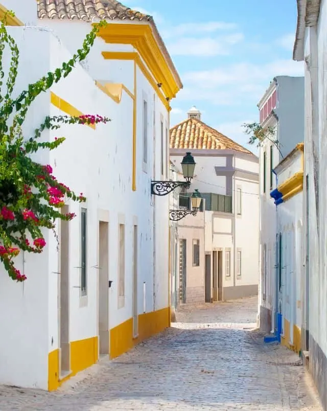 visiting Algarve in December, View down a narrow cobbled street lined with white buildings with gold and blue painted trim on a bright sunny day